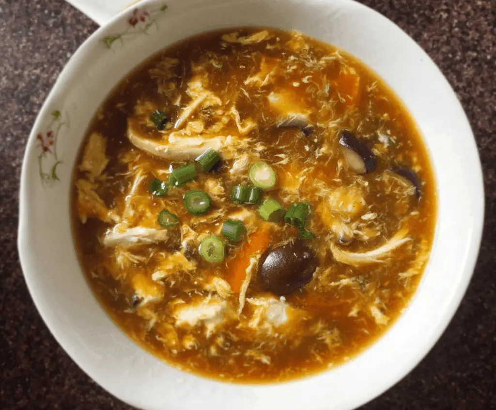 A steaming bowl of Chinese Hot and Sour Soup garnished with sliced mushrooms, tofu, bamboo shoots, and green onions, served in a white ceramic bowl on a wooden table.