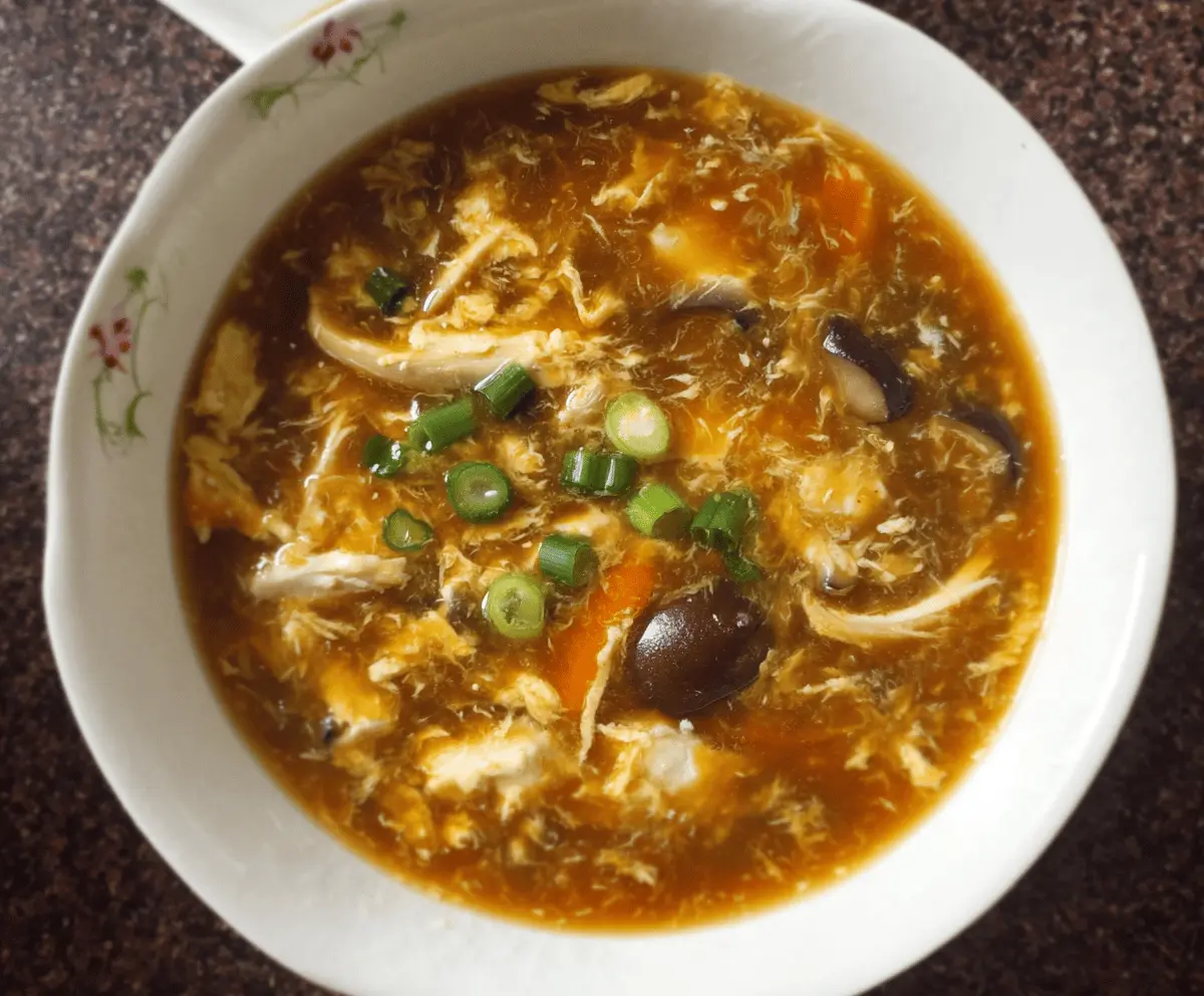 A steaming bowl of Chinese Hot and Sour Soup garnished with sliced mushrooms, tofu, bamboo shoots, and green onions, served in a white ceramic bowl on a wooden table.