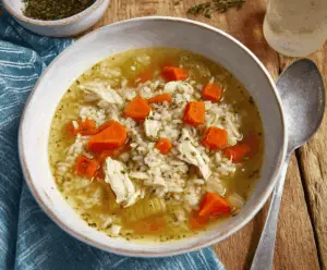 A steaming bowl of homemade chicken and rice soup with shredded chicken, rice, vegetables, and herbs in a clear broth
